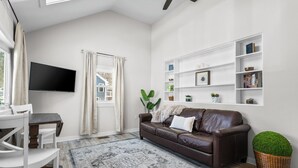 A living room with a brown leather sofa, a flat-screen TV mounted on the wall, a dining table with chairs, and a patterned rug. - The Essex of Gloucester (Gloucester)