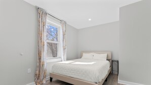 A bedroom with a bed, a window with curtains, a bedside table, and a wall-mounted light. - The Essex of Gloucester (Gloucester)