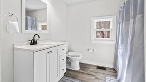 A bathroom with a white vanity, a mirror with lights, a toilet, and a window. - The Essex of Gloucester (Gloucester)