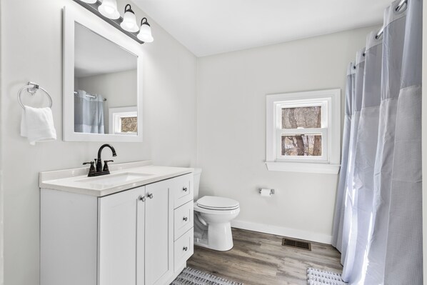 A bathroom with a white vanity, a mirror with lights, a toilet, and a window. - The Essex of Gloucester (Gloucester)