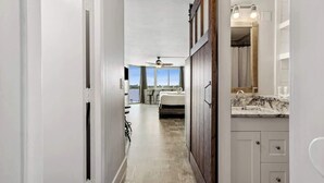 Un pasillo estrecho que conduce a un dormitorio con un ventanal grande, ventilador de techo y un baño con tocador y espejo.