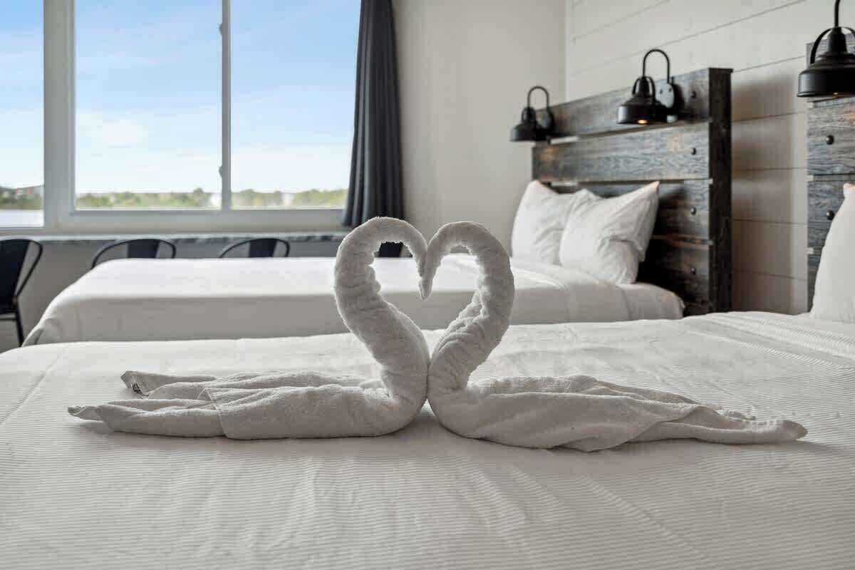 A hotel room with a bed featuring a heart-shaped towel arrangement, a wooden headboard, and a view of the outdoors.