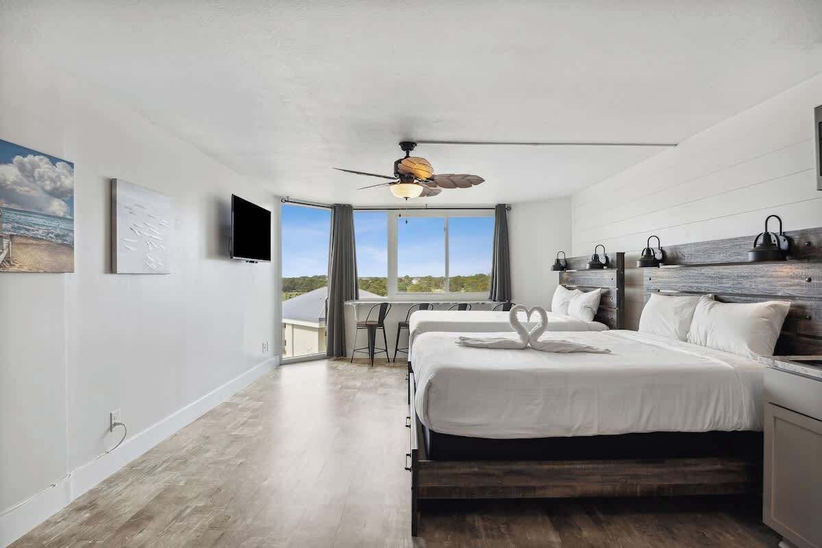 A bedroom with a large bed, a ceiling fan, a desk, and a view of the outdoors.