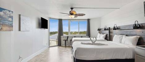A bedroom with a large bed, a ceiling fan, a desk, and a view of the outdoors.