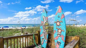 Twee surfplanken steunen tegen een houten hek, met een strand en oceaan op de achtergrond.