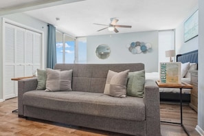 Ein Wohnzimmer mit grauem Sofa, Deckenventilator, einem Willkommensschild und Blick ins Freie durch ein Fenster.