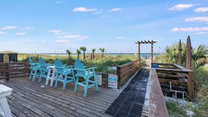 Een houten terras met turquoise Adirondack-stoelen, een houten pergola en uitzicht op het strand.