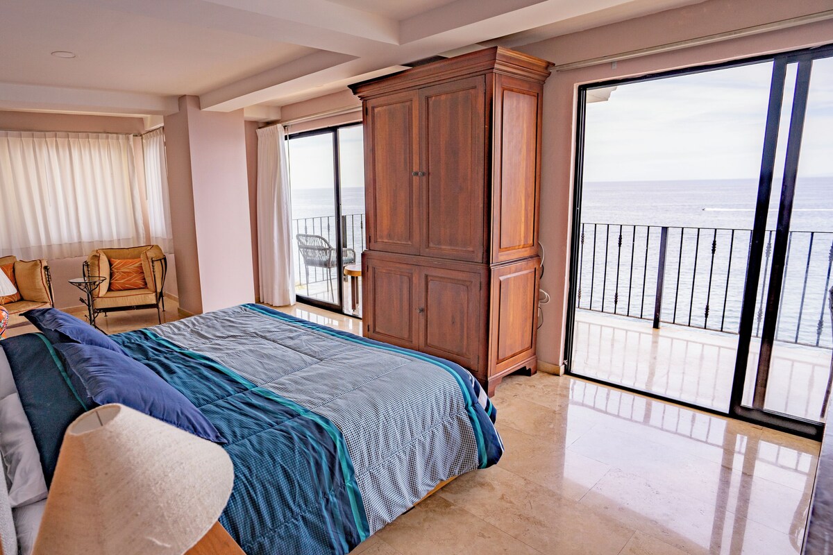 A bedroom with a large bed, a chair, a table, and a view of the sea through a balcony.