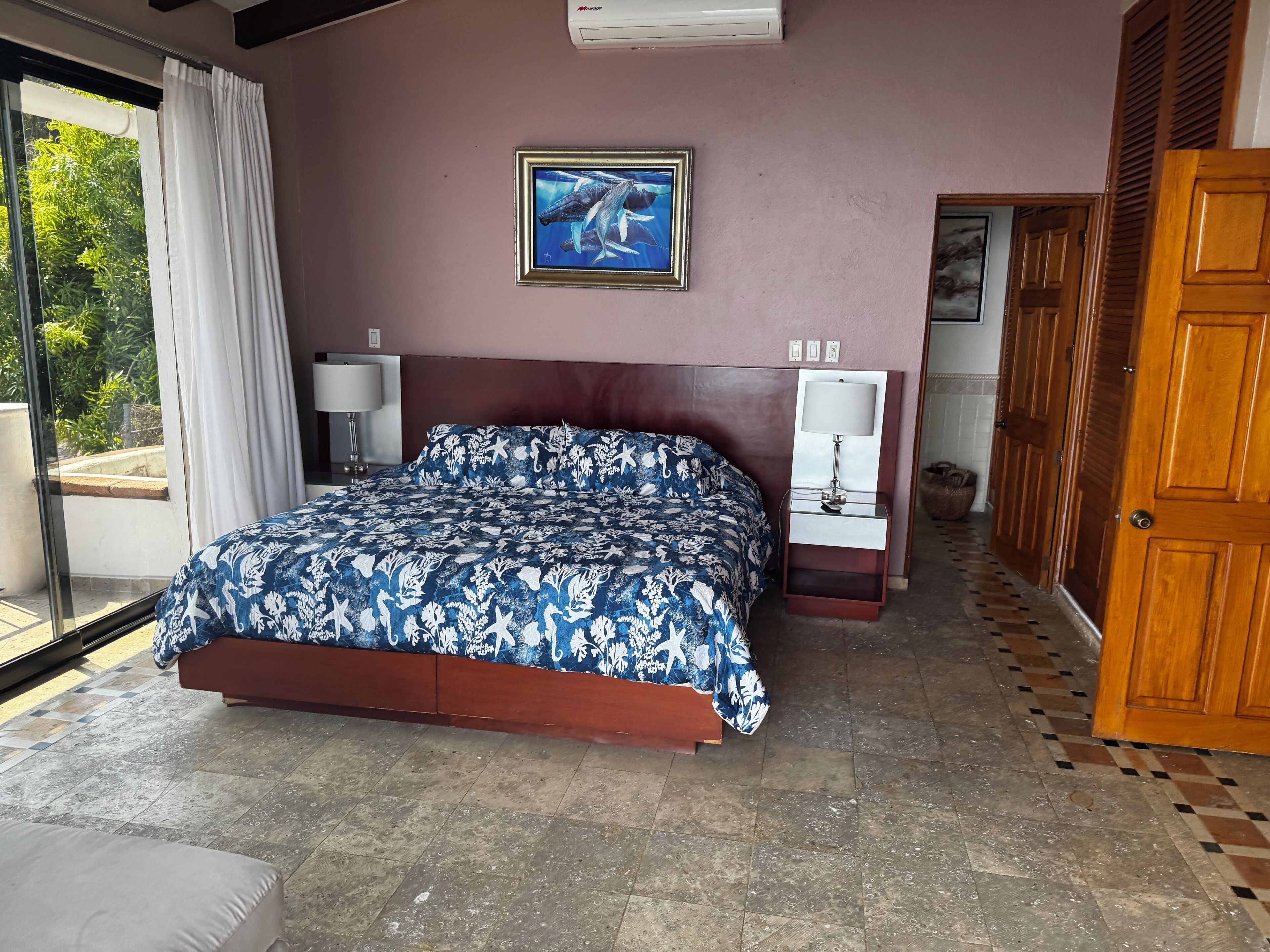 Un dormitorio con cama, mesita de noche, lámpara, un cuadro en la pared y vistas al exterior con vegetación.