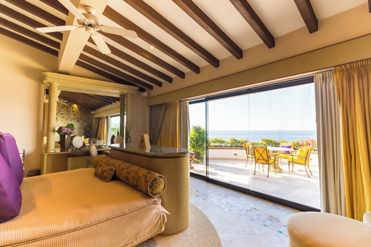 A hotel room with a bed, a desk, a mirror, and a balcony with yellow chairs and a view of the sea.