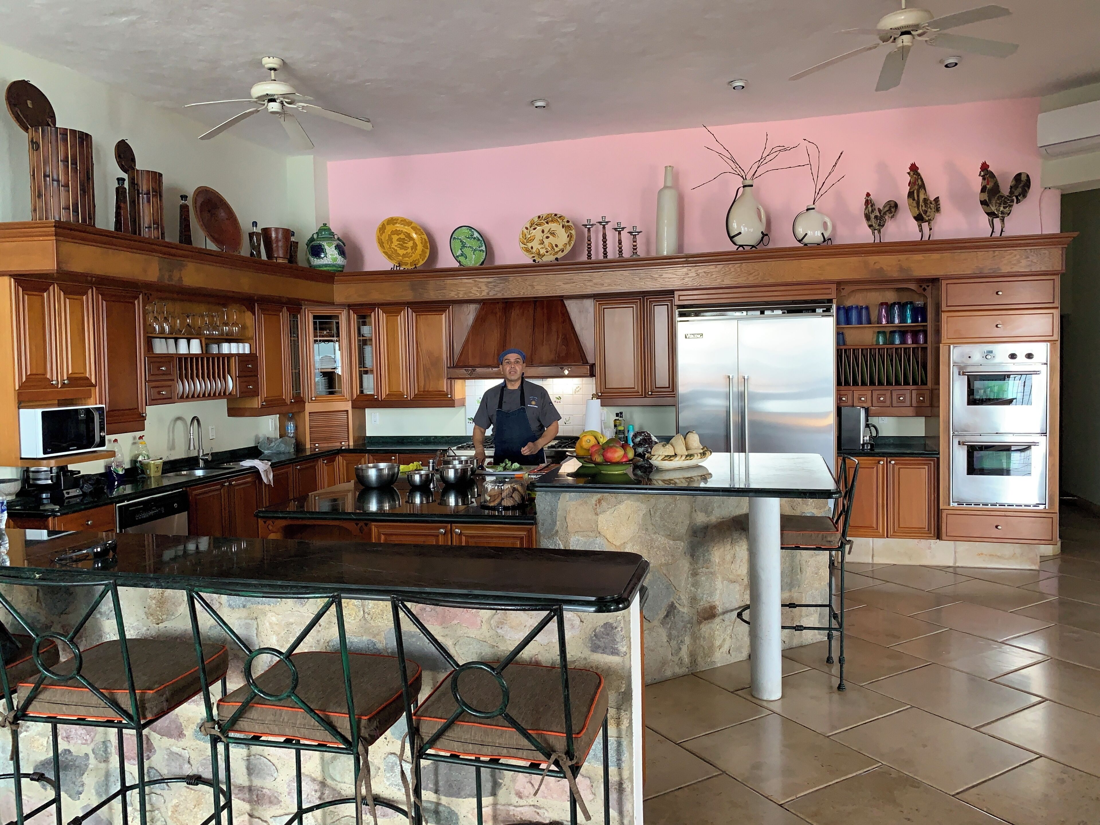 Una cocina con armarios de madera, una isla central, taburetes de bar, un refrigerador y elementos decorativos en la pared.