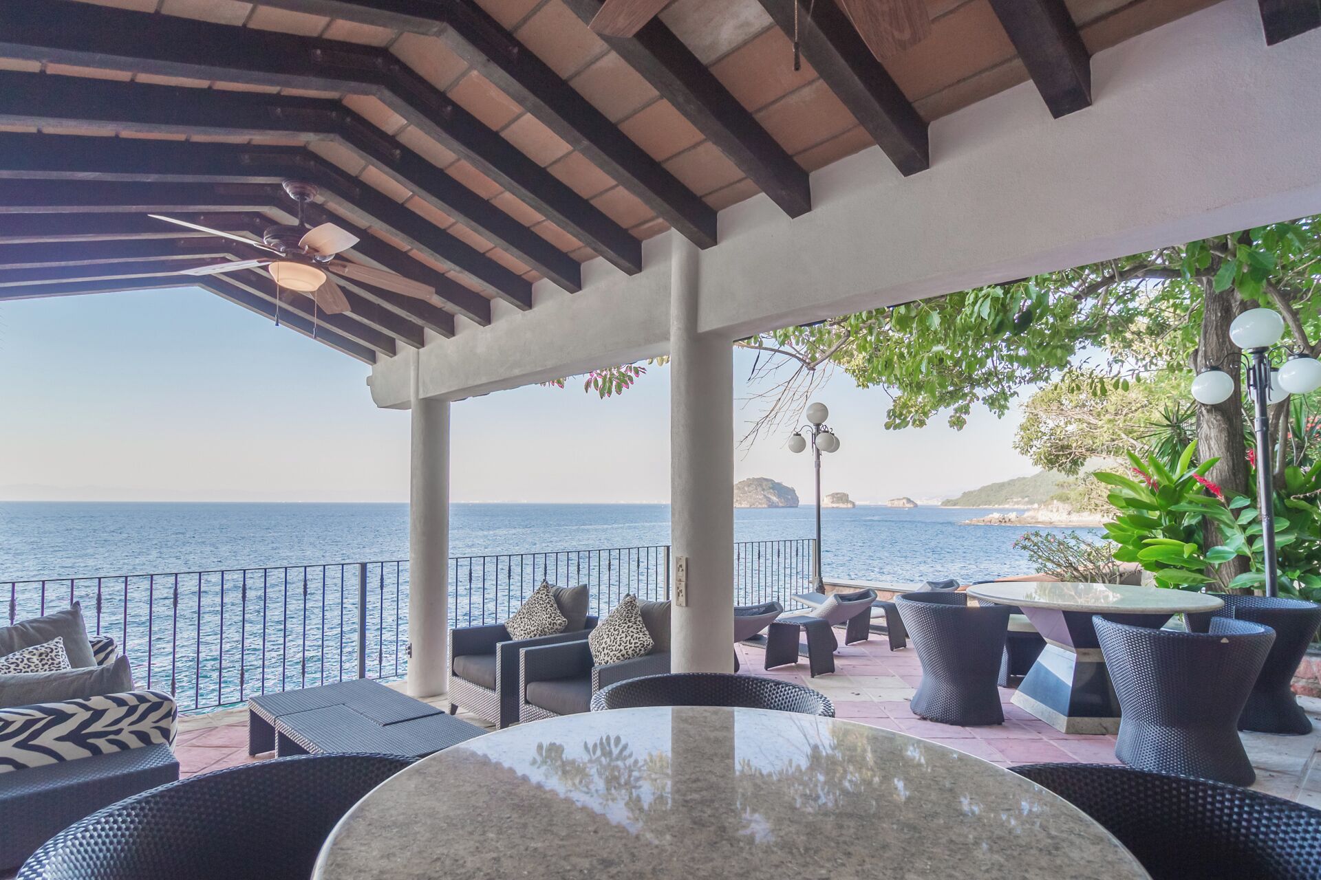A covered outdoor seating area with a view of the sea, featuring wicker furniture and a round table.
