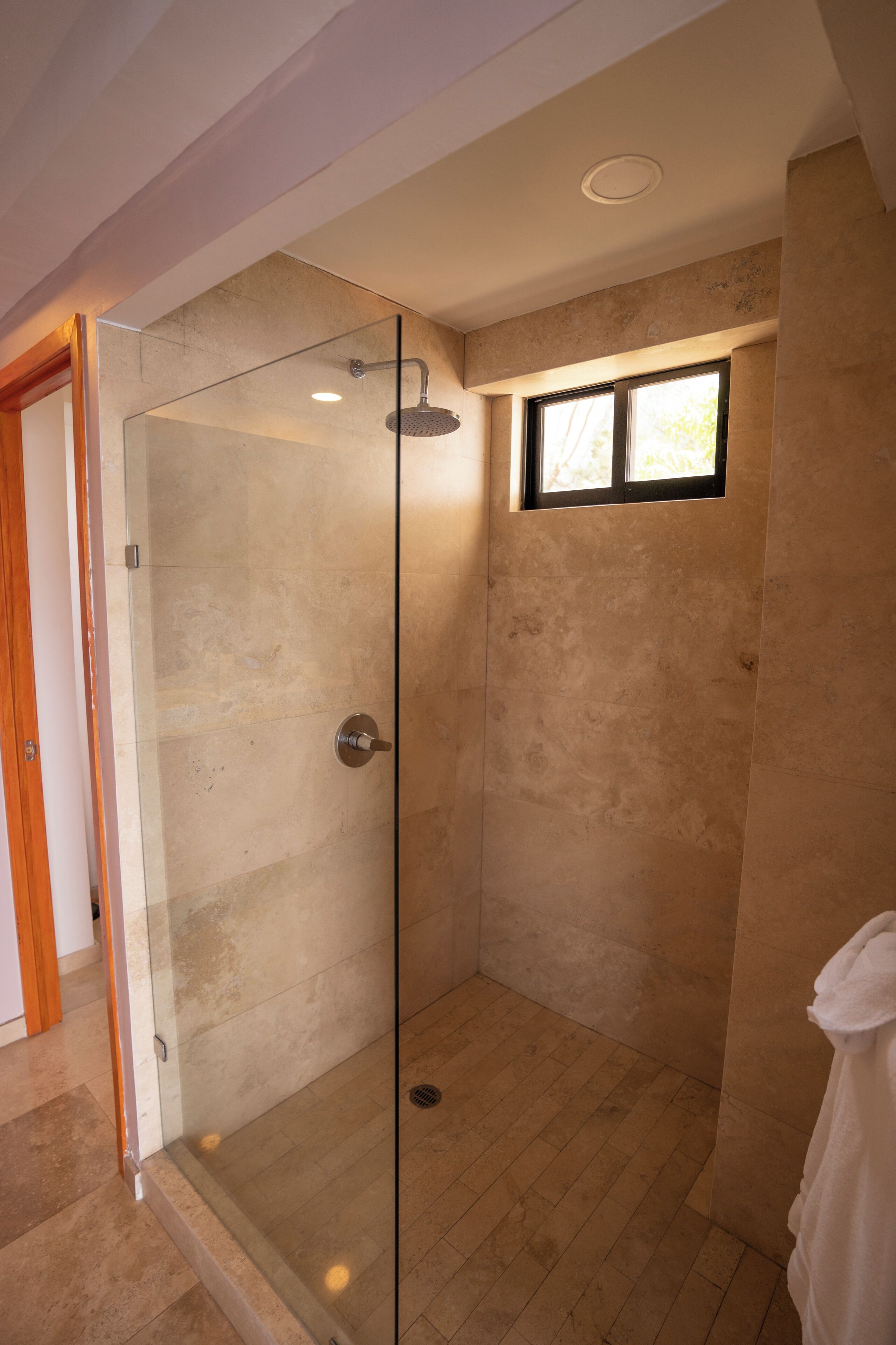 Ducha con mampara de vidrio, cabezal de ducha manual, ventana y una toalla colgada en la pared.