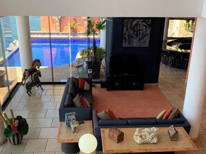 Sala de estar con sofá, mesa de centro y mueble para la televisión. Se ve una piscina a través de puertas de vidrio.