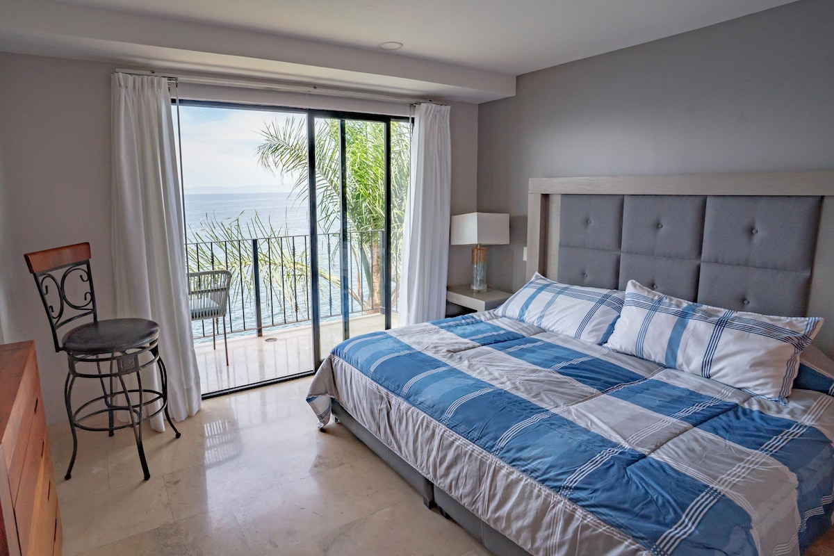 A bedroom with a large bed, a chair, a nightstand, and a view of the ocean through a sliding door.