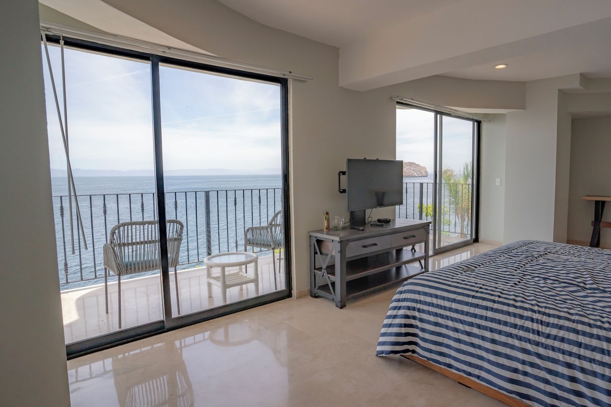 A bedroom with a large bed, a TV, and a balcony with chairs.