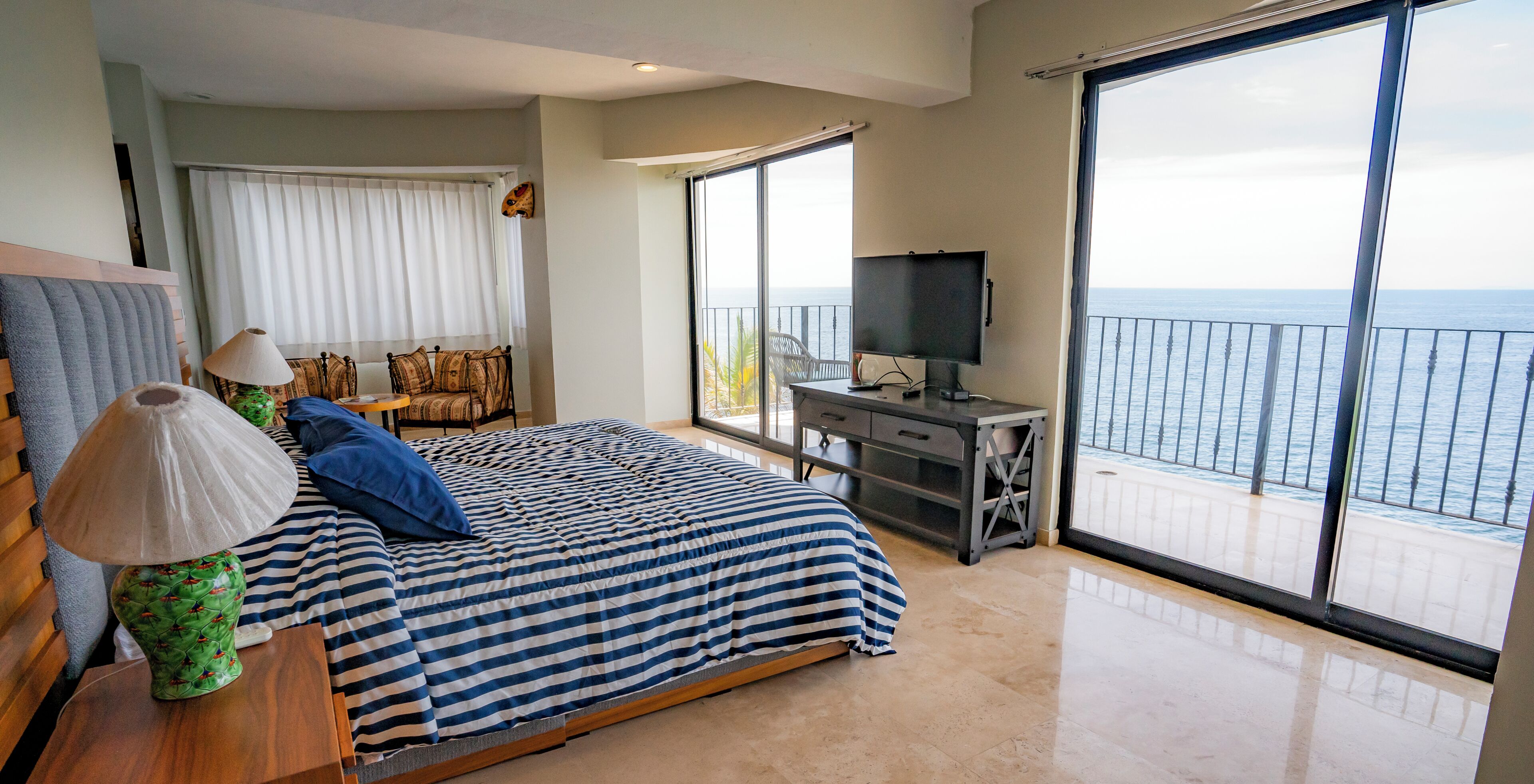 Un dormitorio con cama, escritorio, televisión y balcón con vistas al mar.