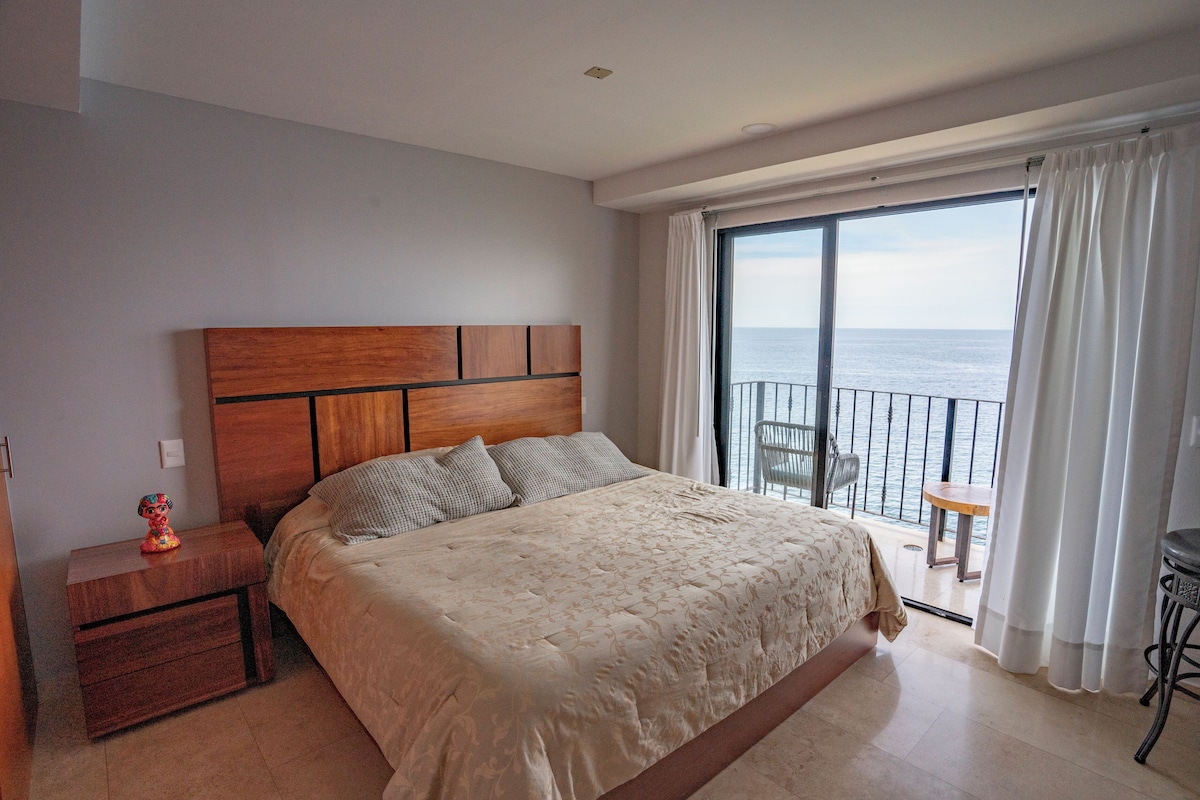 A bedroom with a large bed, a wooden headboard, a nightstand, and a view of the sea through a balcony.
