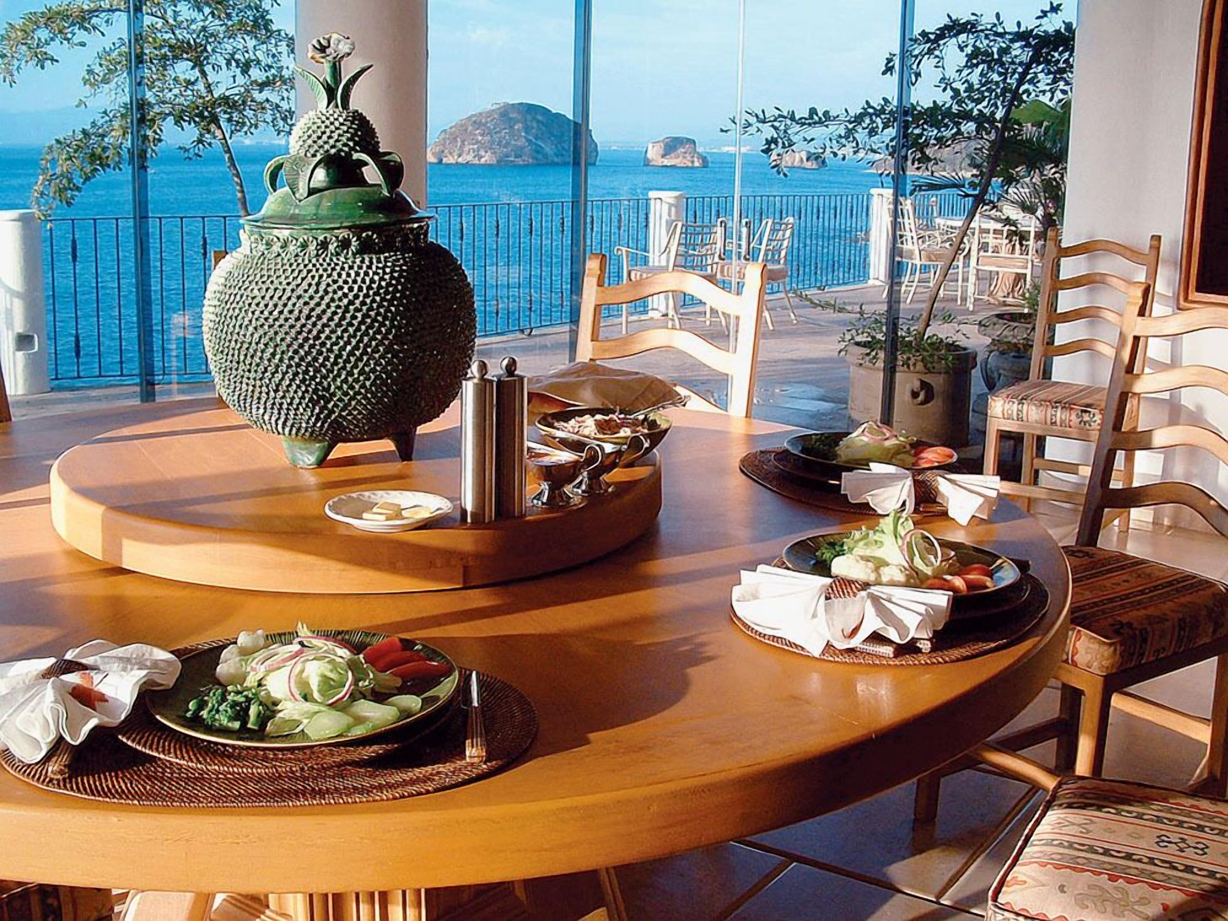 Una mesa de comedor con platos de comida, un jarrón decorativo y sillas en un espacio con vistas al mar.