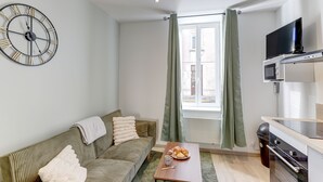 A modern living room with a sofa, a coffee table, a clock, a window with curtains, and a kitchenette with a TV and microwave. - Le Cosy Vert - Proche du Cher (Montlucon)