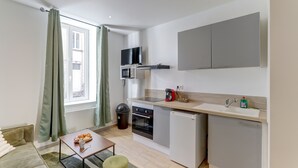 A compact kitchen with a microwave, oven, and sink, a small dining area with a table and chairs, and a window with curtains. - Le Cosy Vert - Proche du Cher (Montlucon)