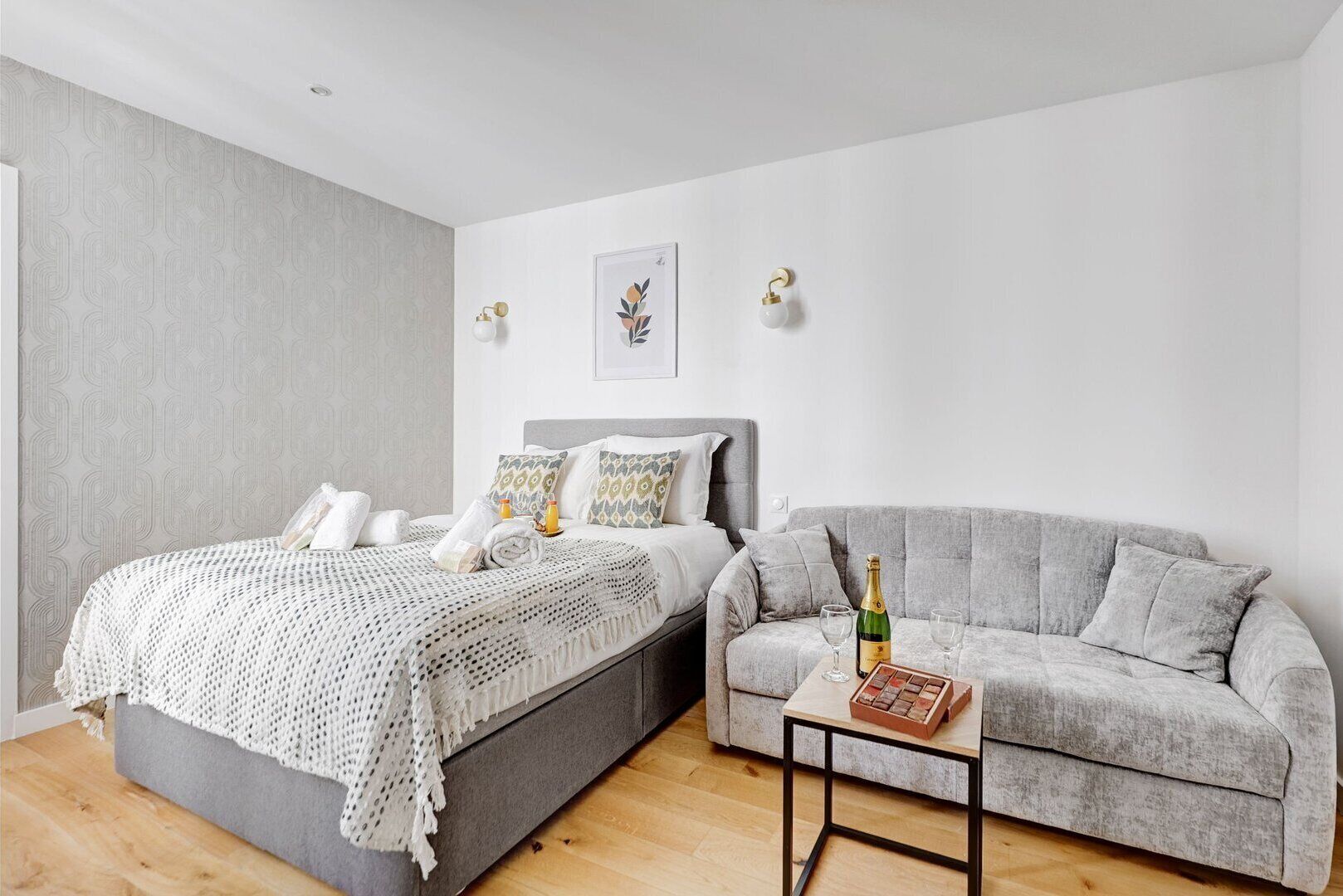 A modern bedroom with a bed, a grey sofa, a small table with a bottle and glasses, and two wall-mounted lights.