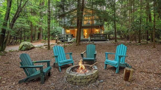 Eine Hütte, umgeben von Bäumen, mit Feuerstelle und vier Adirondack-Stühlen.