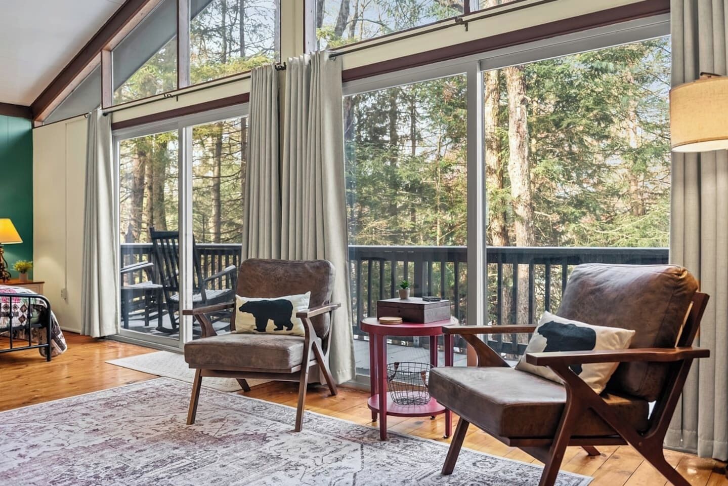 A room with a large window, a wooden floor, a chair with a bear pillow, a small table, and a lamp.
