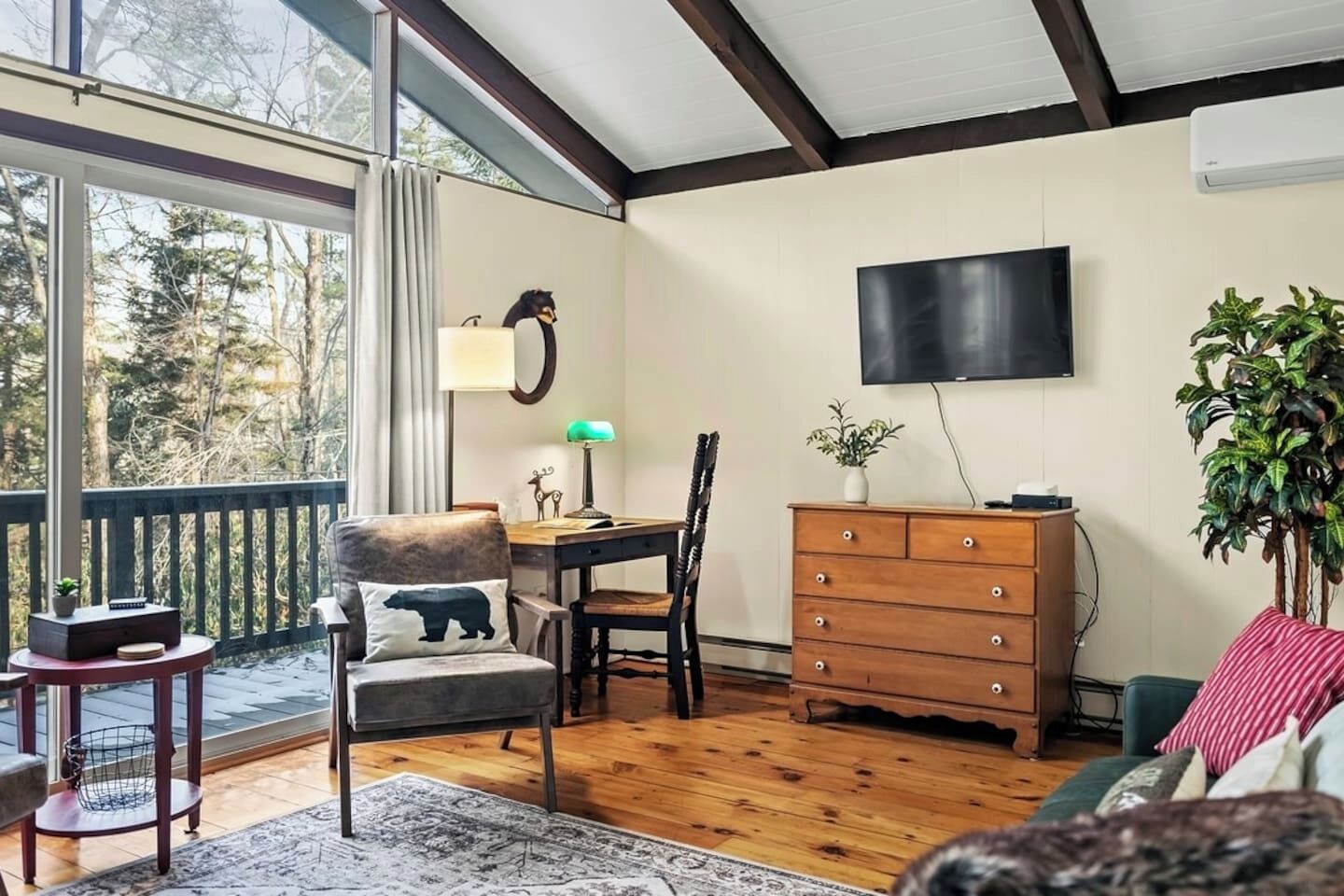 A room with a wooden floor, a desk with a lamp, a chair with a bear pillow, a dresser, a TV on the wall, and a balcony with a railing.