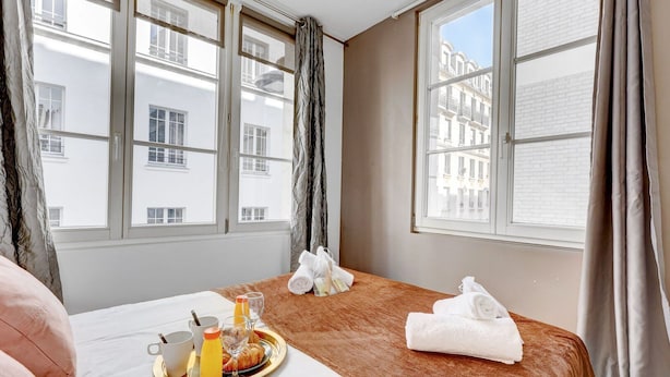 Une chambre d’hôtel avec un lit, un plateau de petit-déjeuner, des serviettes et de grandes fenêtres donnant sur des immeubles.
