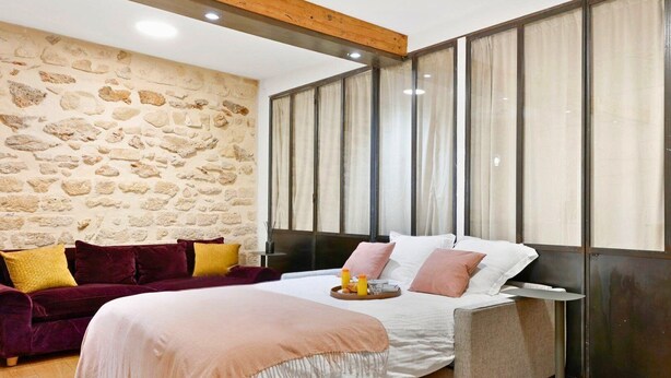 Une chambre avec un mur en pierre, un lit doté d’une tête de lit en bois, un canapé rouge foncé et une table de chevet sur laquelle se trouve un plateau avec divers objets.