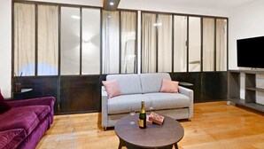 Un salon avec un canapé gris, un canapé violet, une table basse ronde sur laquelle se trouvent une bouteille et une boîte, et un téléviseur posé sur un meuble.