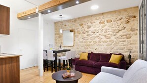 Un salon chaleureux avec un mur en pierre, un canapé violet, un fauteuil gris et une table à manger en bois.