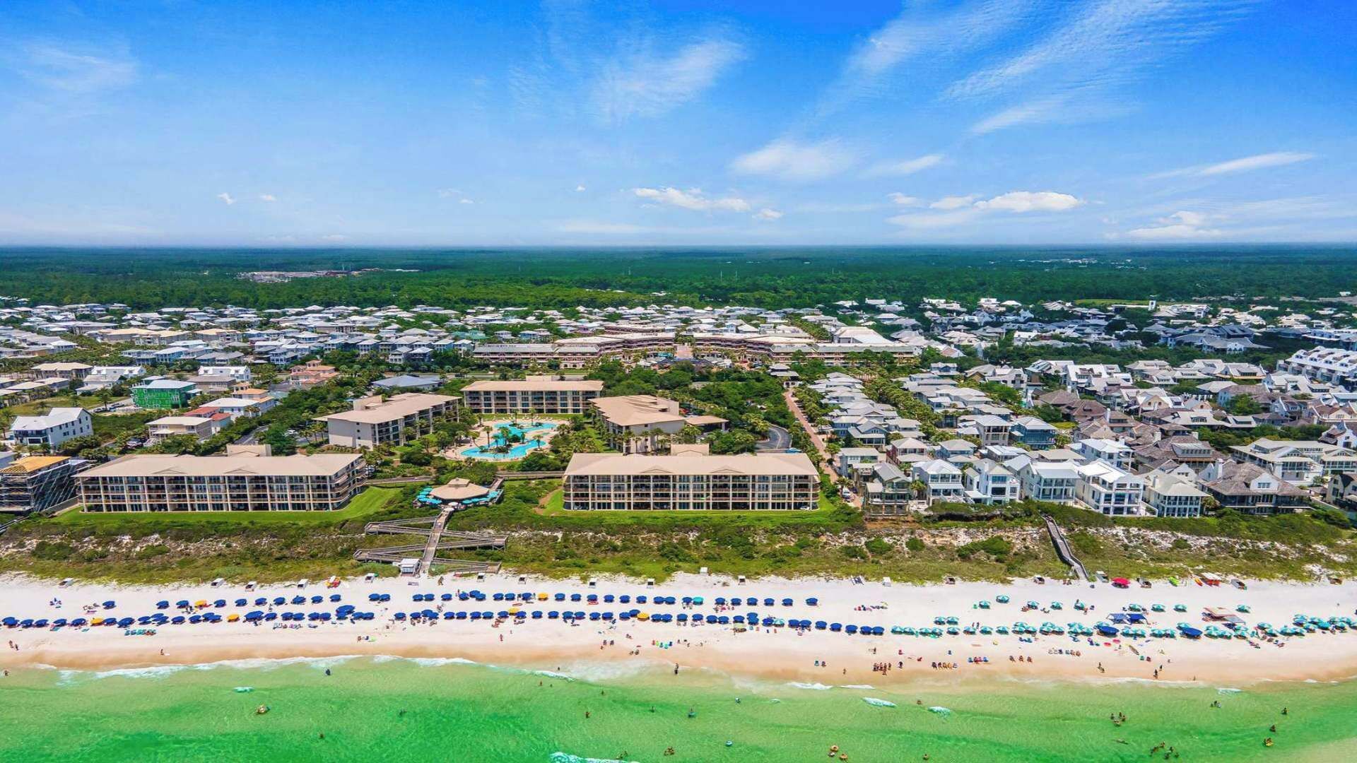 An aerial view of a coastal resort with multiple buildings, a central pool area, and a beach with umbrellas.