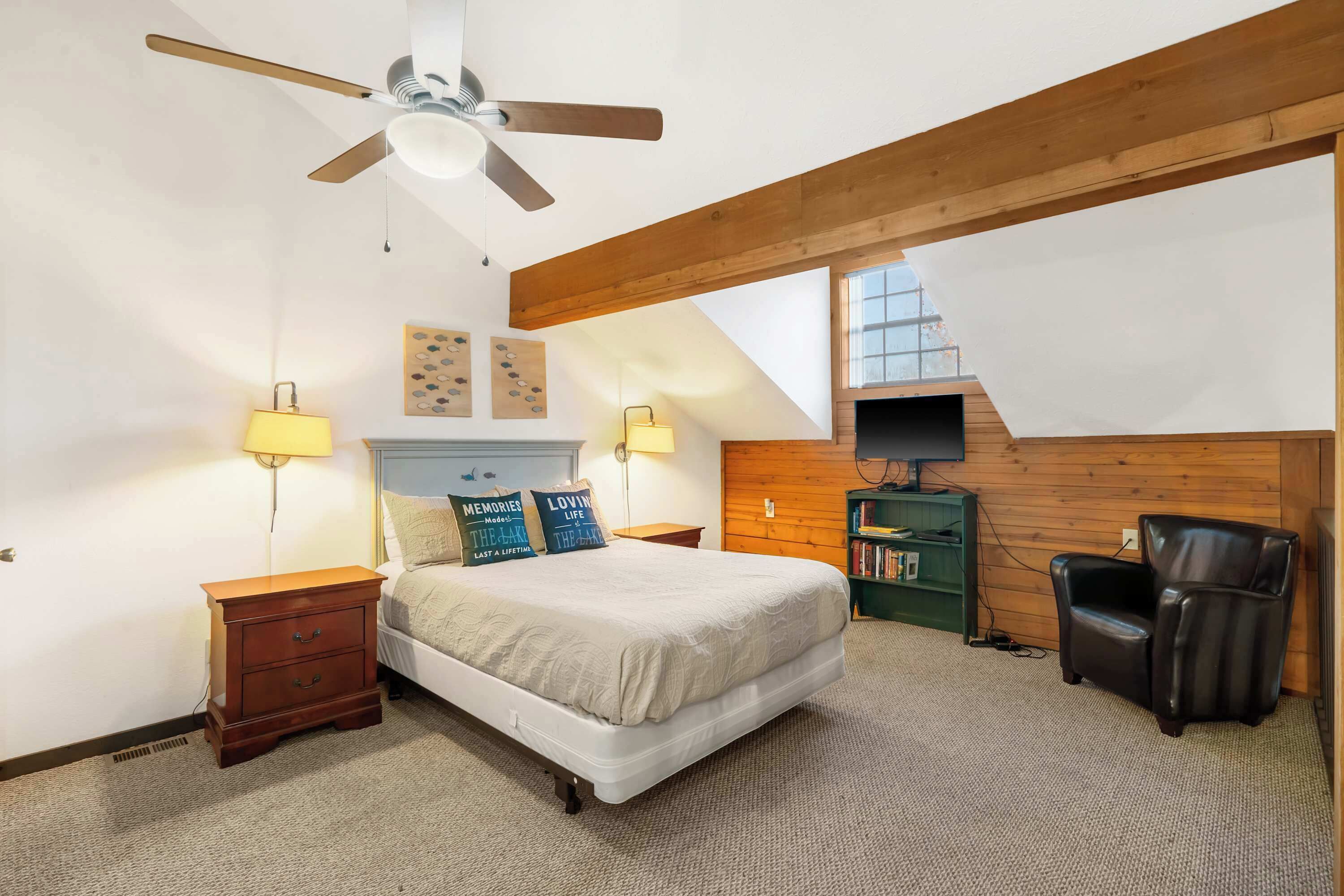 A bedroom with a bed, two bedside lamps, a wooden nightstand, a leather chair, a TV on a wooden shelf, and a ceiling fan.