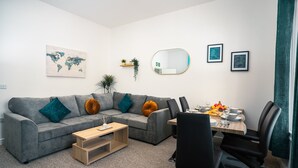 A modern living room with a grey sofa, a wooden coffee table, and a dining area with a table set for two. - Central YeovilStay|Contractors|Free Wi-Fi|Sleeps 8 (Yeovil)