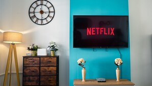 A living room with a teal accent wall, a flat-screen TV displaying "NETFLIX", a wall clock, a wooden cabinet, and a floor lamp. - Central YeovilStay|Contractors|Free Wi-Fi|Sleeps 8 (Yeovil)