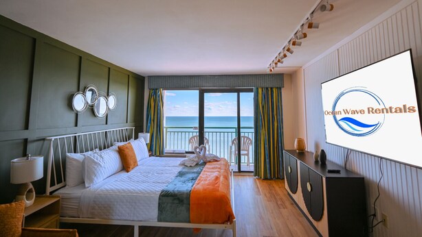 A hotel room with a bed, a TV, a balcony with a view of the sea, and a logo on the wall. - Cozy studio in charming Myrtle Beach with refreshing oceans view (Myrtle Beach)