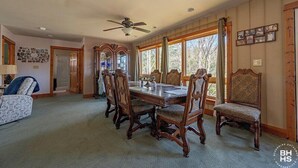 Un comedor con una mesa y sillas de madera, un ventilador de techo y ventanas grandes con cortinas.