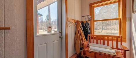 Una habitación con un banco de madera, un perchero con chaquetas, una puerta con ventana y vista a árboles.