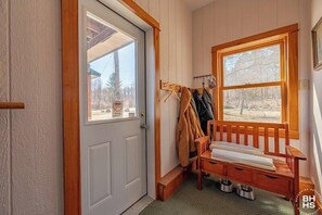 A room with a wooden bench, a coat rack with jackets, a door with a window, and a view of trees.