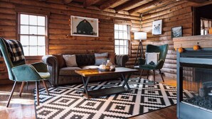 Interior de una cabaña de troncos con un sofá, dos sillones, una mesa de centro y una chimenea.