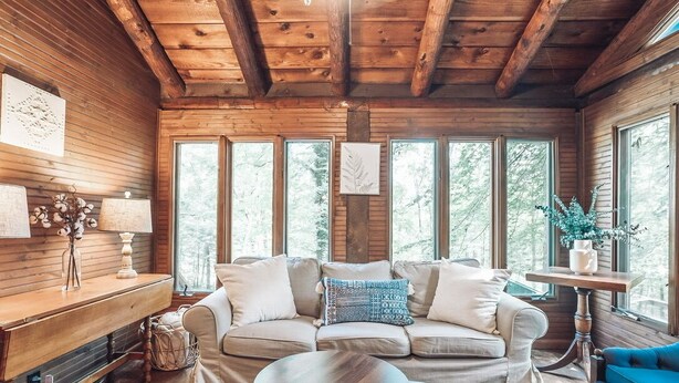 Interior acogedor de una cabaña con techo de madera, un sofá con cojines, una mesita y un ventanal con vistas a árboles.