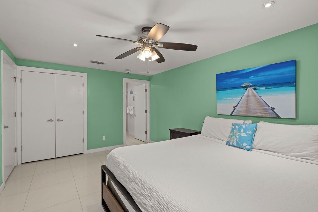 A bedroom with a bed, a ceiling fan, a painting of a beach scene, and a wardrobe.