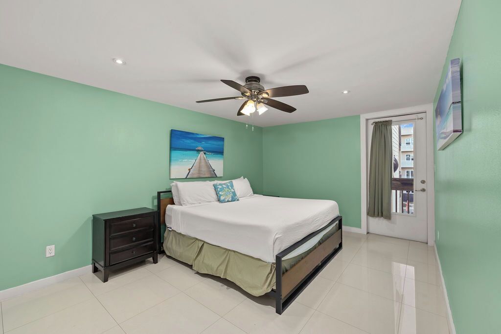 Quarto com cama, mesa de cabeceira, ventilador de teto e pintura de uma cena de praia.
