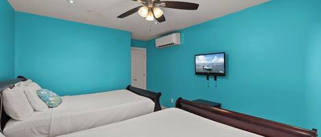 Ein Zimmer mit zwei Betten, einem Deckenventilator, einer Klimaanlage und einem Wand-Fernseher.