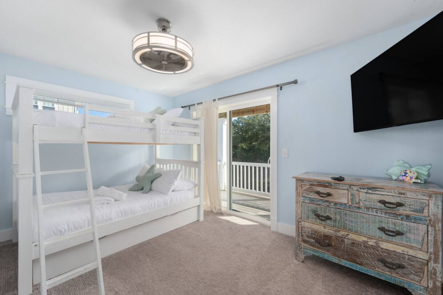 A room with bunk beds, a wooden dresser, a ceiling fan, and a TV mounted on the wall.