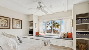 Een slaapkamer met een bed, een vensterbank met uitzicht op de oceaan, een plafondventilator en opbergplanken.