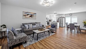 A modern living room with a grey sofa, patterned armchairs, a coffee table, and a dining area with a table and chairs.