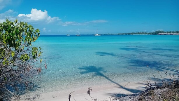 Une plage bordée d’une eau turquoise et limpide, quelques voiliers, et une côte lointaine parsemée de bâtiments.